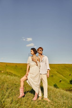 asian woman in wedding dress and cowboy boots near redhead groom in sunglasses in countryside
