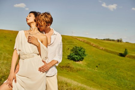 redhead man in sunglasses embracing elegant asian bride on green meadow, tranquil rustic wedding