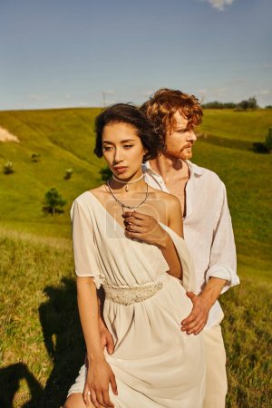sensual asian woman in white wedding dress near young redhead groom in picturesque rustic setting