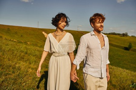 Téléchargez les photos : Joyeux jeunes mariés multiethniques tenant la main et marchant dans la campagne avec un paysage pittoresque - en image libre de droit