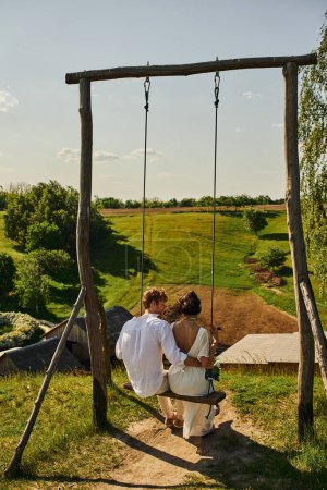 rustic wedding, back view of young newlyweds on swing in countryside with green scenic landscape