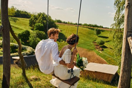 alegre pelirroja novio balanceo con novia en vestido blanco en el campo con pintoresco paisaje