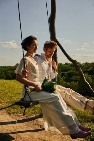 happy asian bride with bouquet swinging with redhead groom in scenic countryside, rustic wedding
