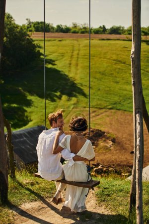 Téléchargez les photos : Marié rousse excité embrassant mariée en robe blanche tout en s'amusant sur swing dans un cadre rural - en image libre de droit