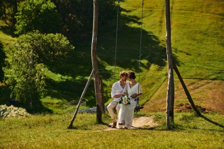 Foto de Feliz momento de pareja multiétnica recién casada en traje de estilo boho en swing en el paisaje - Imagen libre de derechos