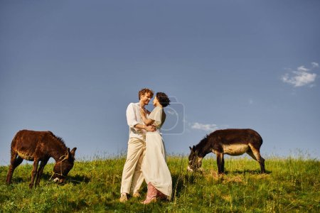 happy multiethnic just married couple embracing near grazing donkeys, idyllic rural setting
