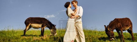 feliz interracial recién casados traje blanco abrazando cerca de burros pastando en el prado, bandera