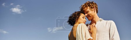 happy redhead man embracing young asian bride under blue sky, tranquil rural wedding, banner