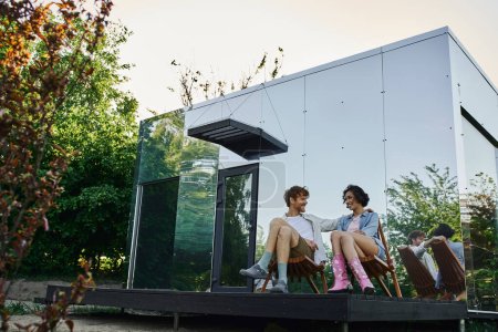 happy multiethnic couple sitting on chairs and looking at each other near modern glass house