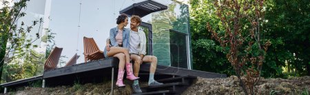 Téléchargez les photos : Joyeux couple multiethnique assis sur le porche de la maison d'herbe moderne et souriant à l'autre, bannière - en image libre de droit