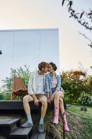 multiethnic couple in stylish casual attire kissing on porch of glass house in countryside