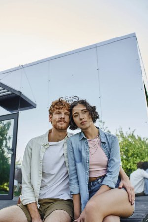 Téléchargez les photos : Couple multiethnique rêveur souriant et regardant loin près de la maison de verre dans la campagne, tranquillité - en image libre de droit