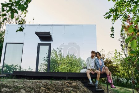 cheerful interracial couple sitting on porch of modern glass house in countryside setting