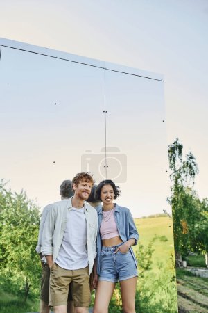 cheerful and stylish multiethnic couple looking away near mirror wall of glass house in countryside