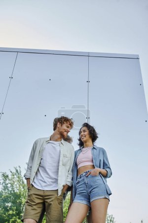 Téléchargez les photos : Couple interracial avec les mains dans les poches souriant à l'autre près du mur miroir de la maison en verre - en image libre de droit