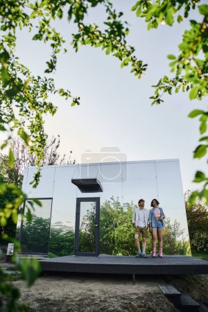 junges gemischtrassiges Paar in stilvoller Freizeitkleidung, das auf der Veranda eines Glashauses im Grünen steht
