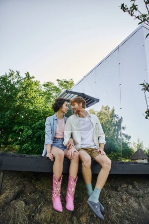 alegre pareja multiétnica sentada en el porche cerca de la casa de cristal y mirándose, felicidad