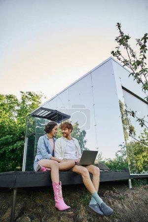 glückliches gemischtrassiges Paar mit Laptop sitzt auf der Veranda in der Nähe von modernen Glashaus auf dem Land