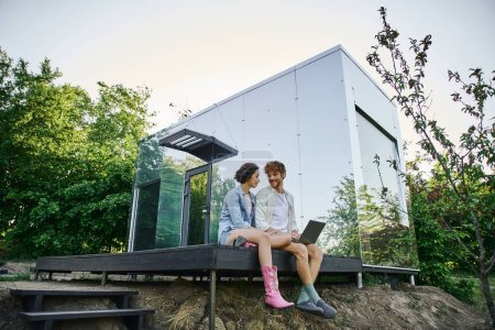 joven pareja multiétnica sentada en el porche con portátil cerca de la casa de cristal moderna en el campo