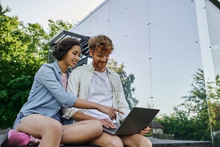 Téléchargez les photos : Jeune femme asiatique pointant vers ordinateur portable près de rousse petit ami sur le porche de la maison en verre dans la campagne - en image libre de droit