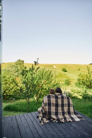 back view of young couple sitting on wooden porch under plaid blanket and enjoying scenic landscape