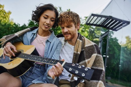 young asian woman playing acoustic guitar to redhead  boyfriend near glass house in countryside