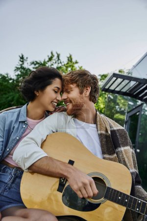 fröhlicher rothaariger Mann spielt Akustikgitarre zu junger asiatischer Freundin in der Nähe von Glashaus auf dem Land