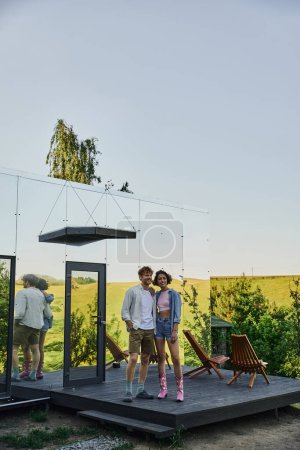 full length of happy interracial couple looking away on porch of modern glass house in countryside
