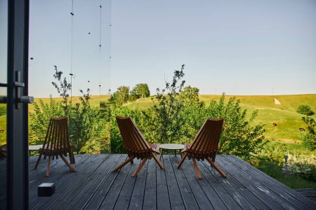 sillas de madera y mesa de centro en el porche cerca de la moderna casa de cristal con vista al paisaje escénico