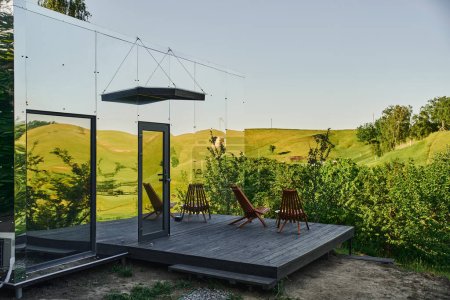 Foto de Casa de cristal ecológica con sillas en el porche de madera y vista al pintoresco paisaje rural - Imagen libre de derechos