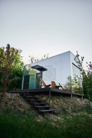 modern eco-friendly glass house with wooden stairs and chair on porch, tranquil countryside setting