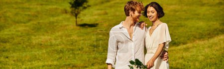joyful redhead groom embracing asian bride with wedding bouquet in scenic countryside, banner