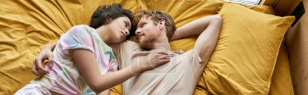 vista superior de la pareja interracial acostada en la cama amarilla y sonriéndose en casa, pancarta