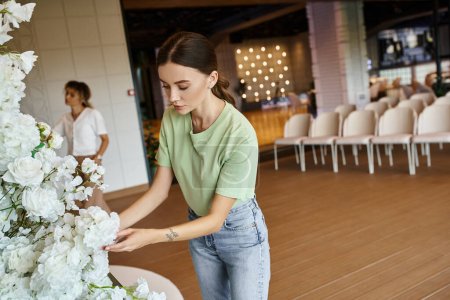 junge tätowierte Frau arbeitet mit blühender floraler Komposition in Veranstaltungshalle neben verschwommenem Kollegen