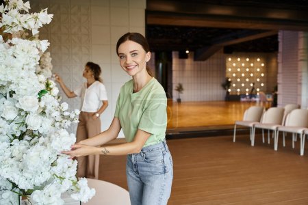 Photo for Happy decorator looking at camera while arranging floral design in event hall near blurred colleague - Royalty Free Image