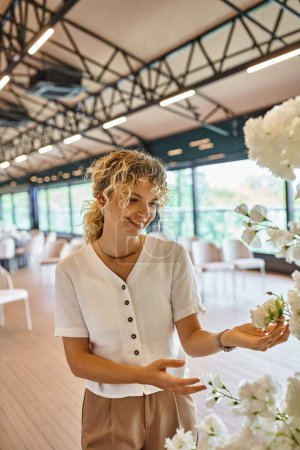 joyful blonde woman with wavy hair smiling near white flowers in event hall, creative florist