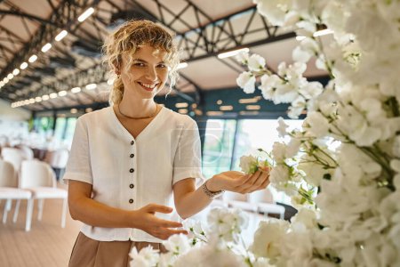 blonde woman with wavy hair smiling near festive floral composition in event hall, creative florist
