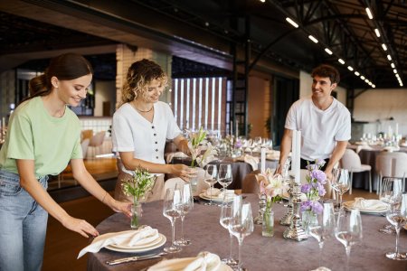 Téléchargez les photos : Équipe joyeuse organiser table festive et placer des fleurs près de la vaisselle dans la salle d'événements - en image libre de droit