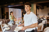 smiling event manager with plates and napkins looking at camera near colleagues and festive table tote bag #675846272