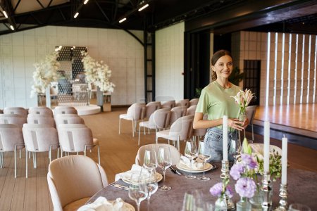 Téléchargez les photos : Joyeux fleuriste tenant vase avec des fleurs près de la table avec vaisselle de fête et des bougies dans la salle des événements - en image libre de droit