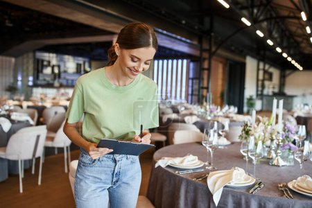 joyful event manager writing notes on clipboard near tables with festive setting in banquet hall