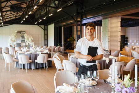 joyous decorator with clipboard looking at camera near festive tables in modern banquet hall