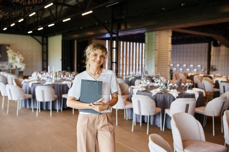 smiling blonde event manager with clipboard looking at camera in banquet hall with decorated tables
