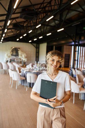 Photo for Cheerful event manager with clipboard looking at camera near decorated tables in banquet hall - Royalty Free Image