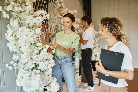 glücklicher Florist zeigt auf weiße florale nahe Teamleitung mit Klemmbrett in der Veranstaltungshalle, kreative Arbeit