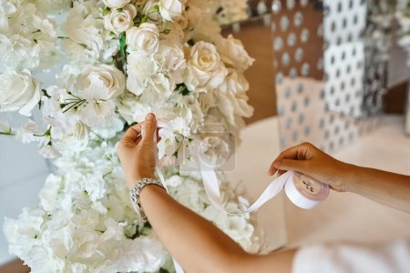 vista recortada de decorador sosteniendo cinta blanca cerca de flores en flor en sala de eventos, configuración de banquete