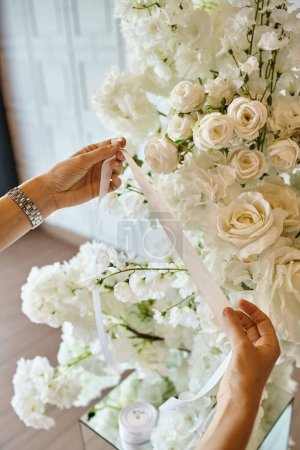 partial view of decorator holding white ribbon near blooming flowers in banquet hall, event setup