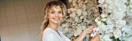 cheerful florist looking at camera while working with white floral decor in event hall, banner