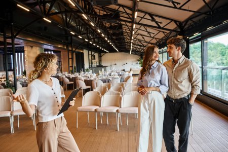 happy couple looking at each other near event manager with clipboard showing modern banquet hall