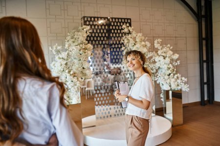 smiling decorator with clipboard showing white floral decor to enamored couple in banquet hall